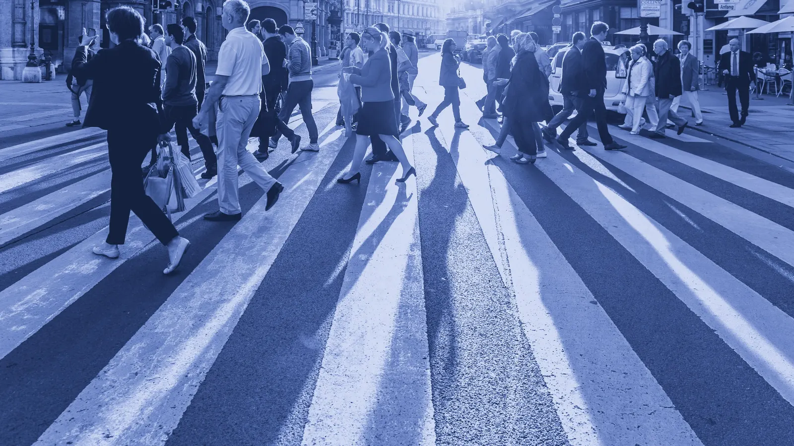 Multidão atravessando uma faixa de pedestres em uma avenida movimentada, em contraluz.