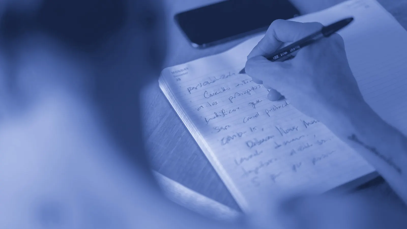 Mão anotando a caneta em um caderno pautado, com um celular apoiado ao lado na mesa.