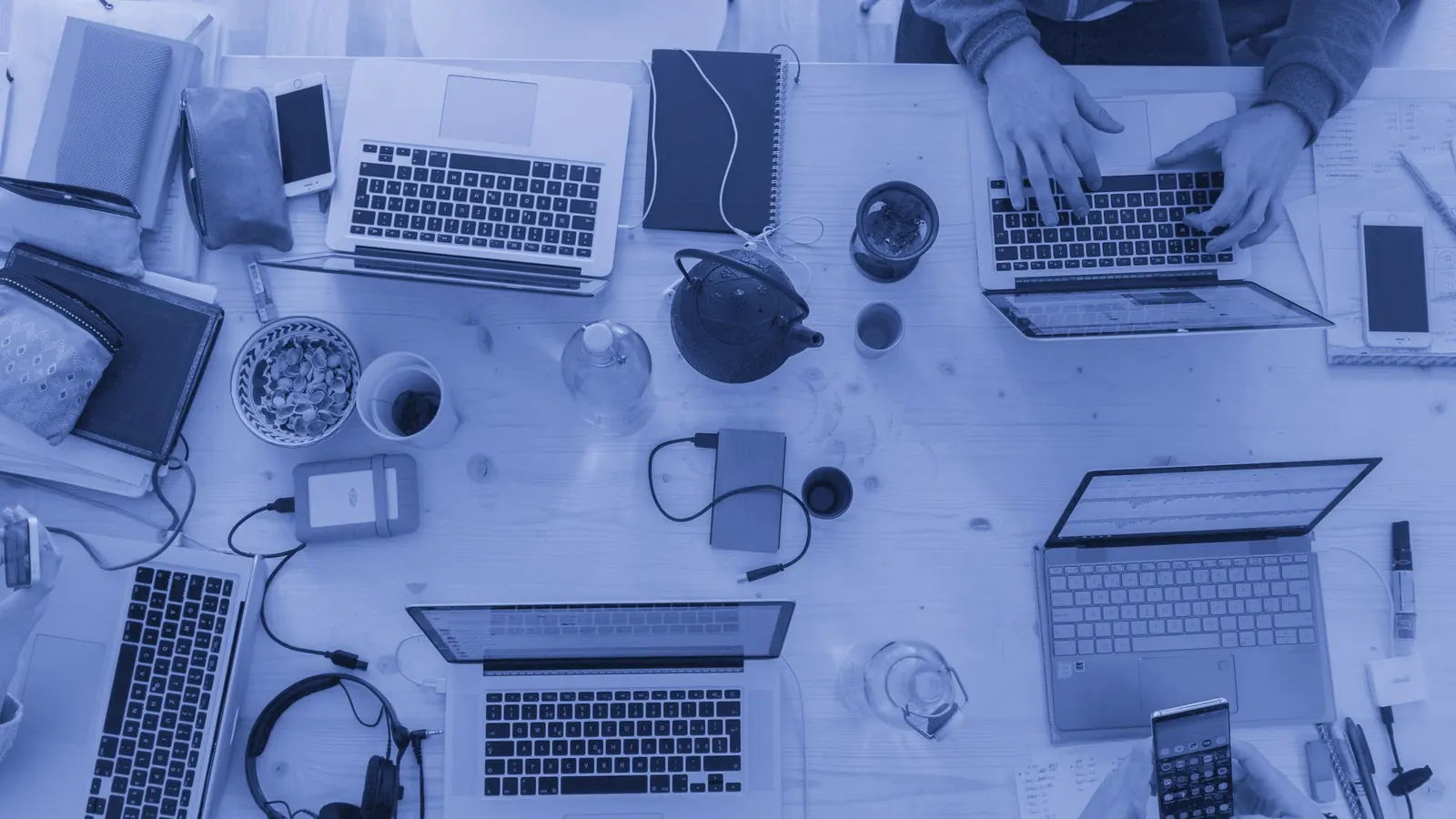 Vista de cima de uma mesa de trabalho com quatro notebooks abertos, cadernos e mãos digitando.